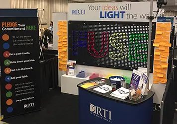 Booth with "RTI" sign displaying "FUSE", surrounded by post-it notes, pamphlets, and a "PLEDGE" board.