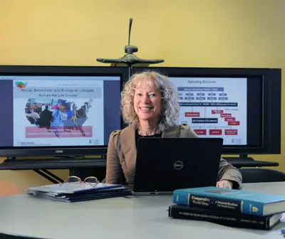 A woman sits at a desk with a laptop, smiling, in front of two presentation screens.